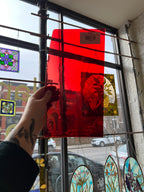 Person holding a red transparent piece of stained glass in front of a blurred sunny window. 