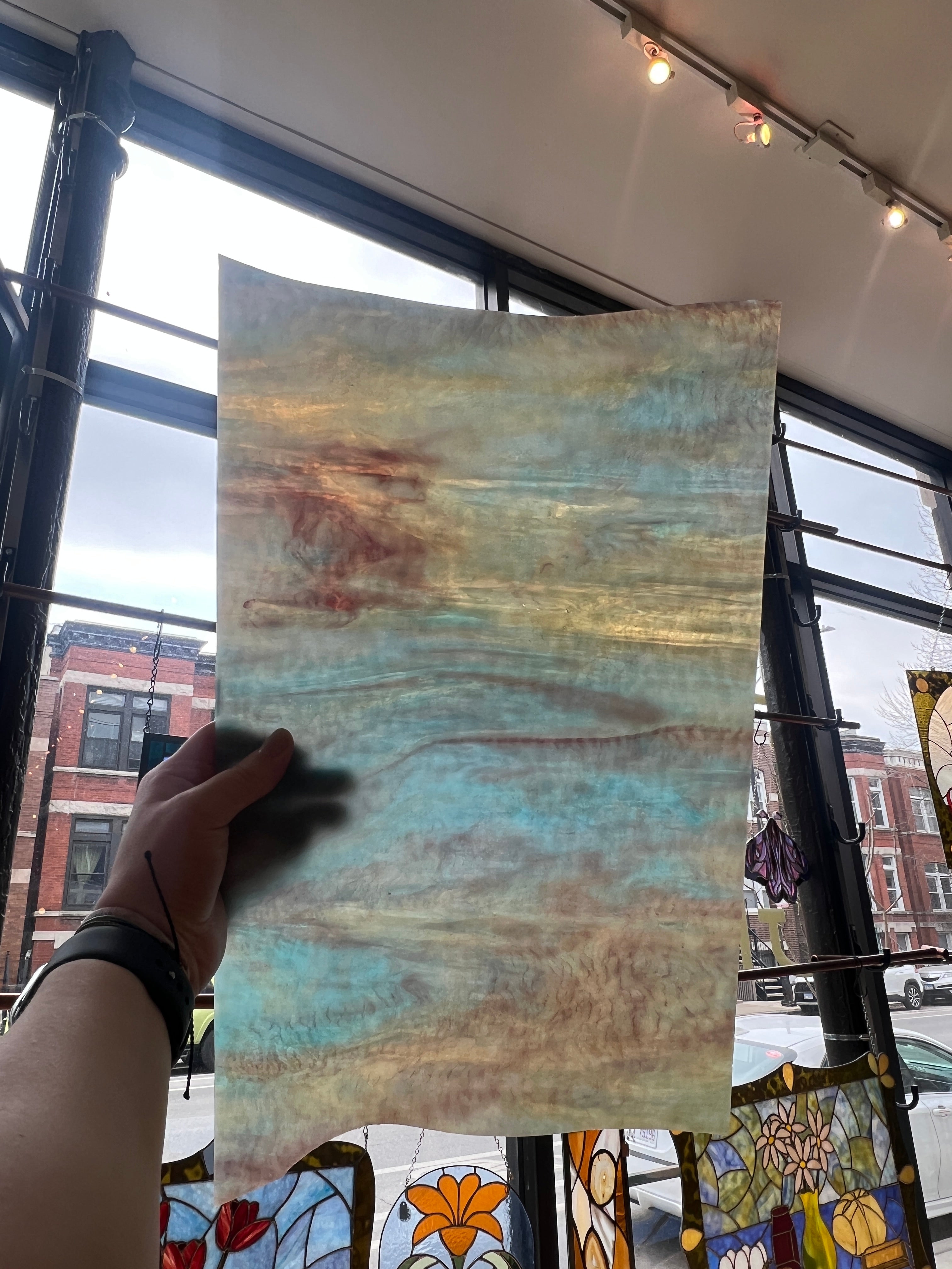 Hand holding a piece of stained glass with a watercolor-like pattern in an indoor setting with stained glass windows.