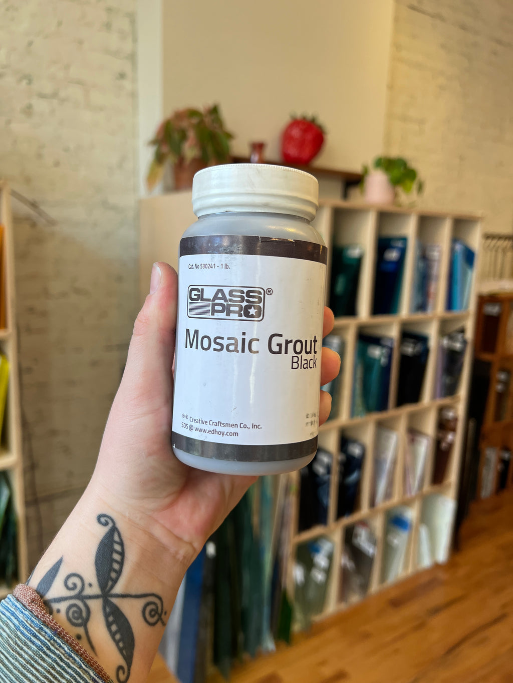 Hand holding a jar of Mosaic Grout Black by Glass Pro with a shelf  in the background.