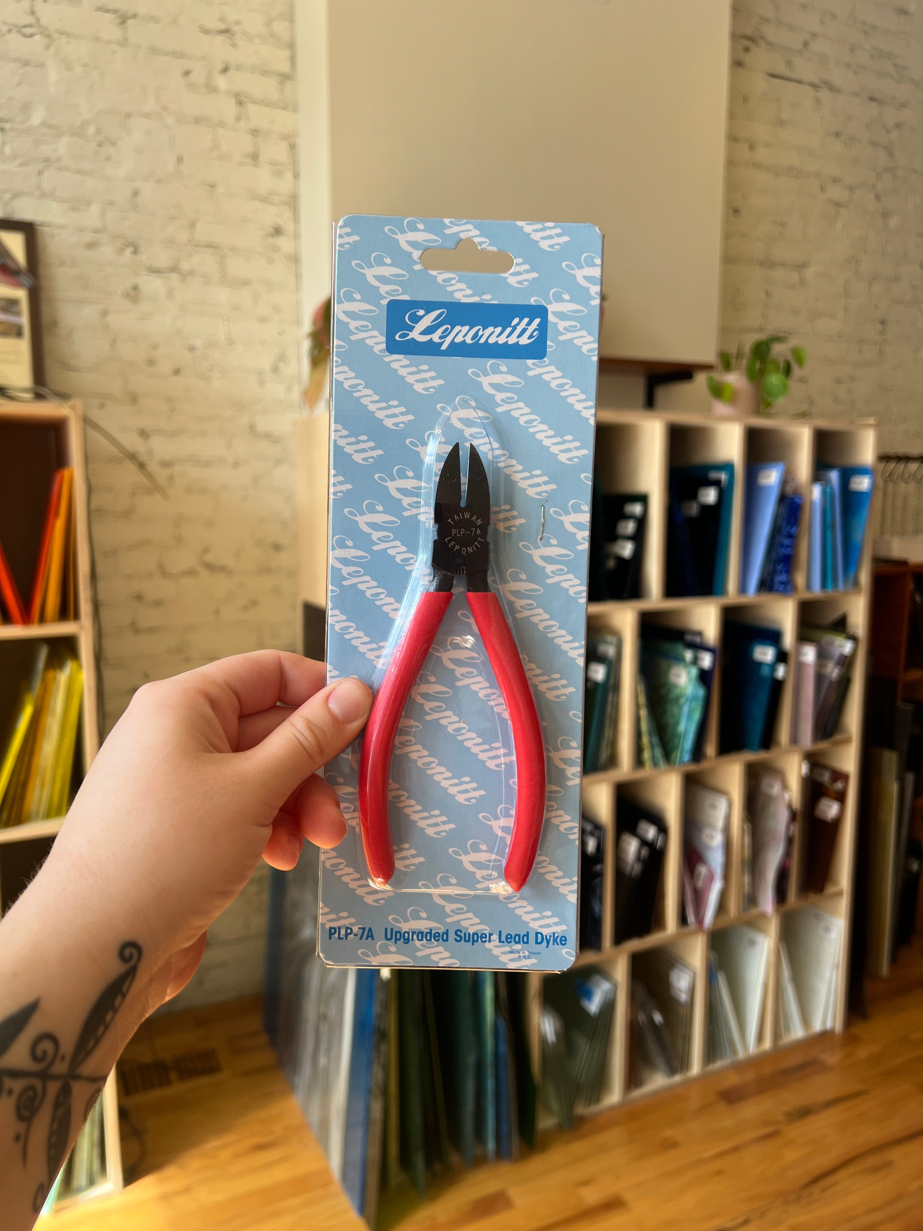 Hand holding a pair of red pliers on a blue card, against a background of shelves.