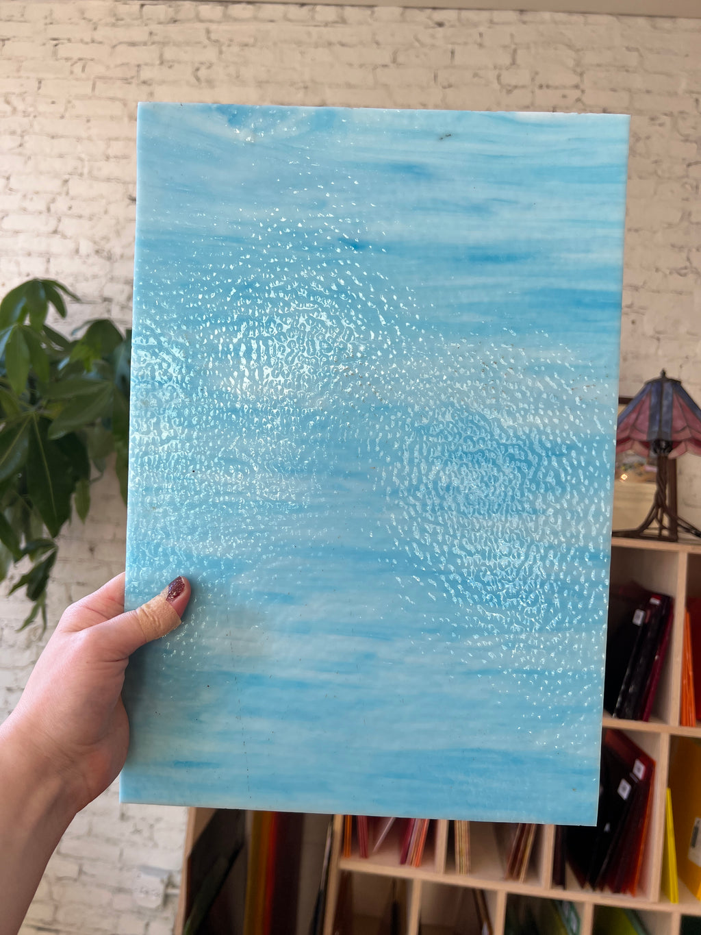 Hand holding a blue textured piece of stained glass against a blurred indoor background
