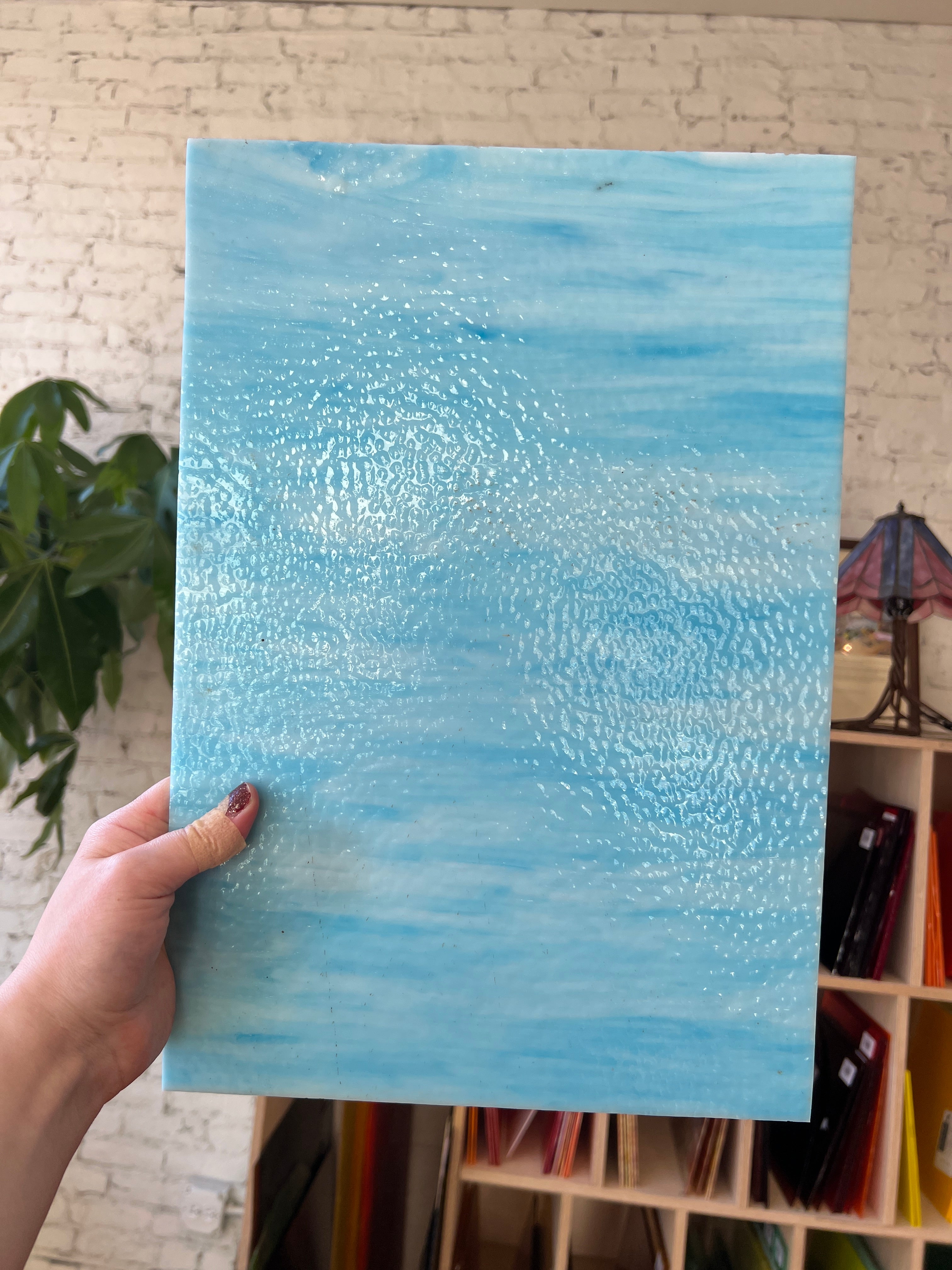 Hand holding a blue textured piece of stained glass against a blurred indoor background