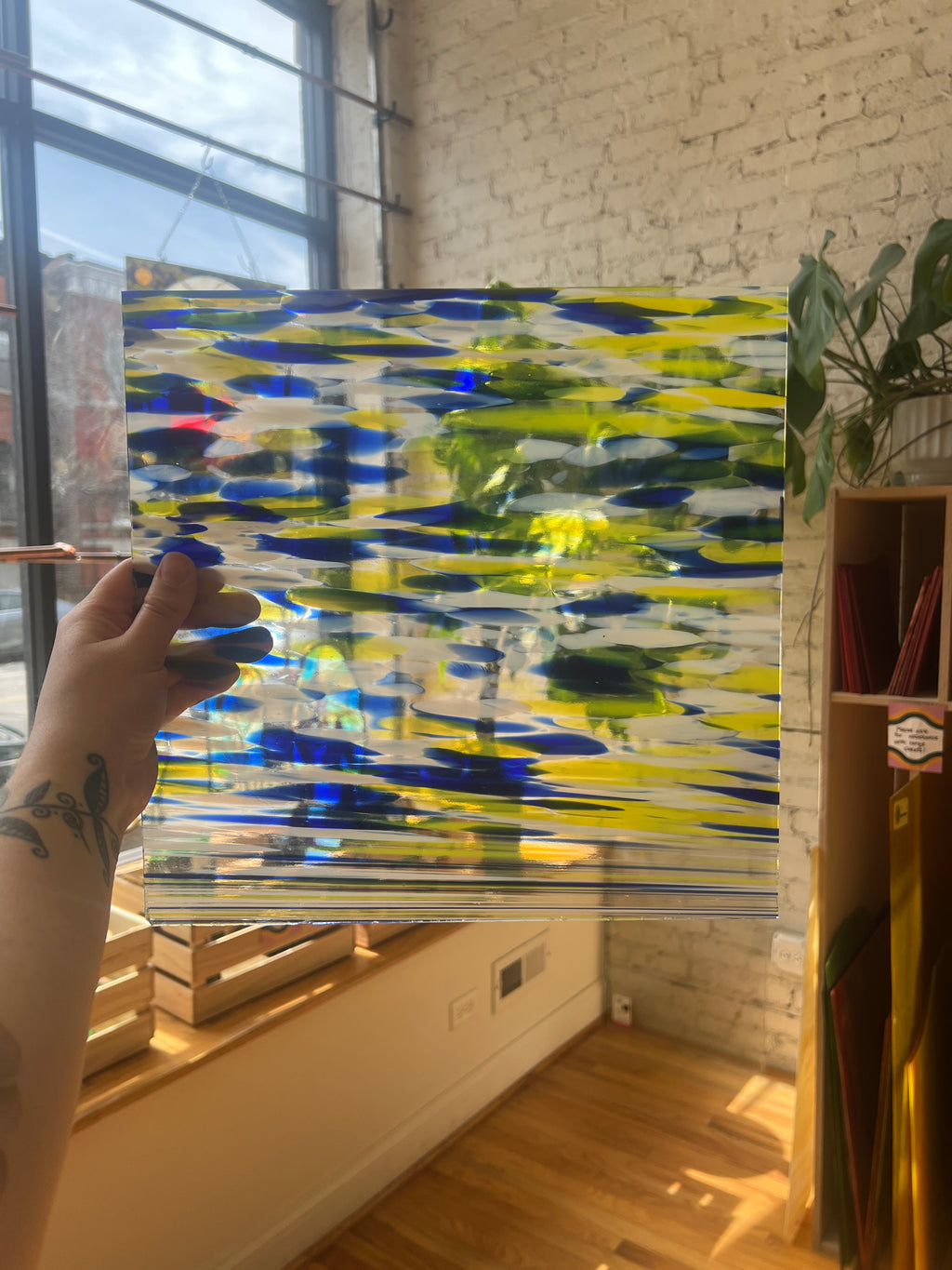 Hand holding a colorful piece of stained glass in a room with a window and plant.