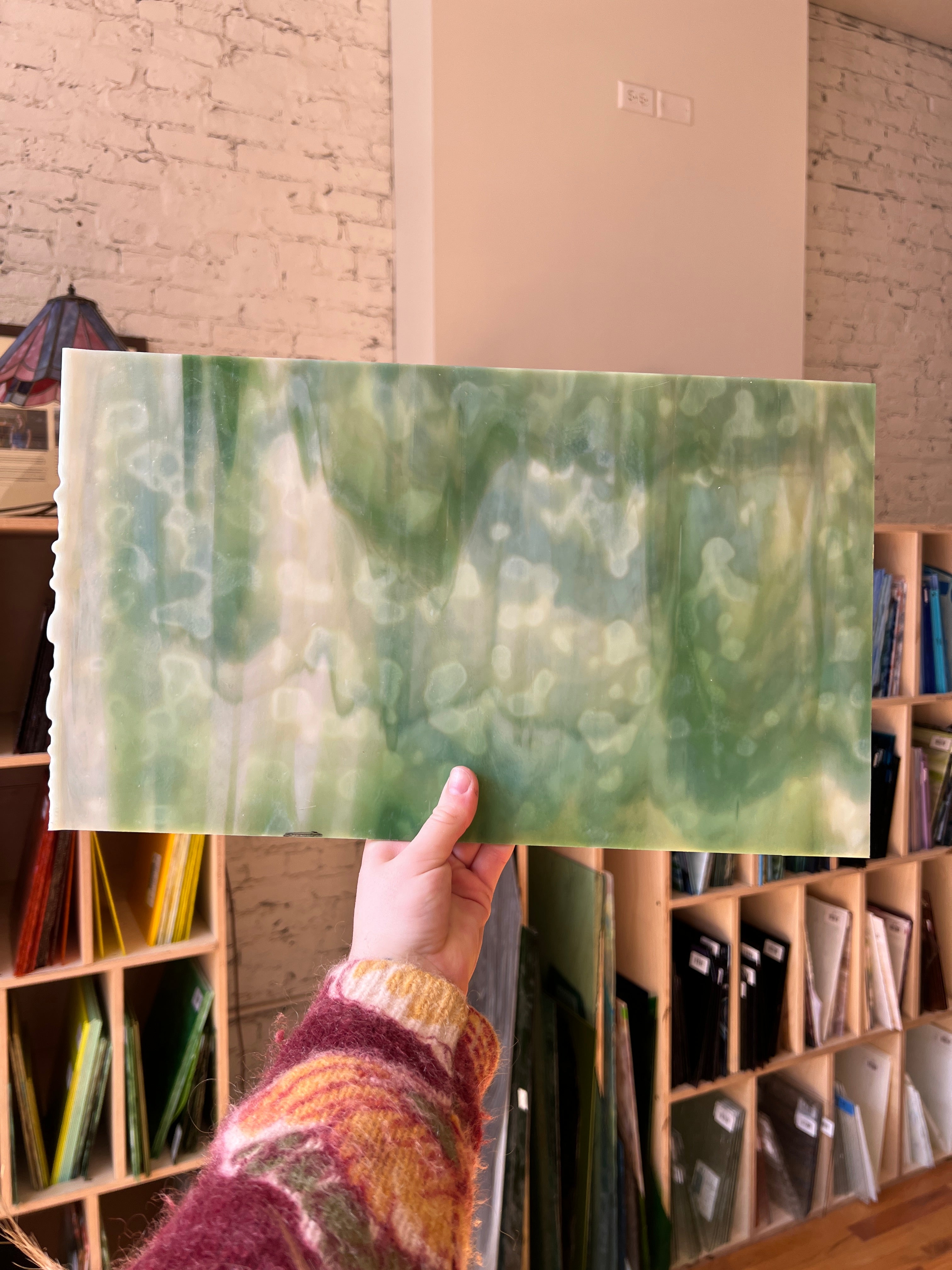 Hand holding a green marbled paper sheet of stained glass with a background of shelves filled with similar items.
