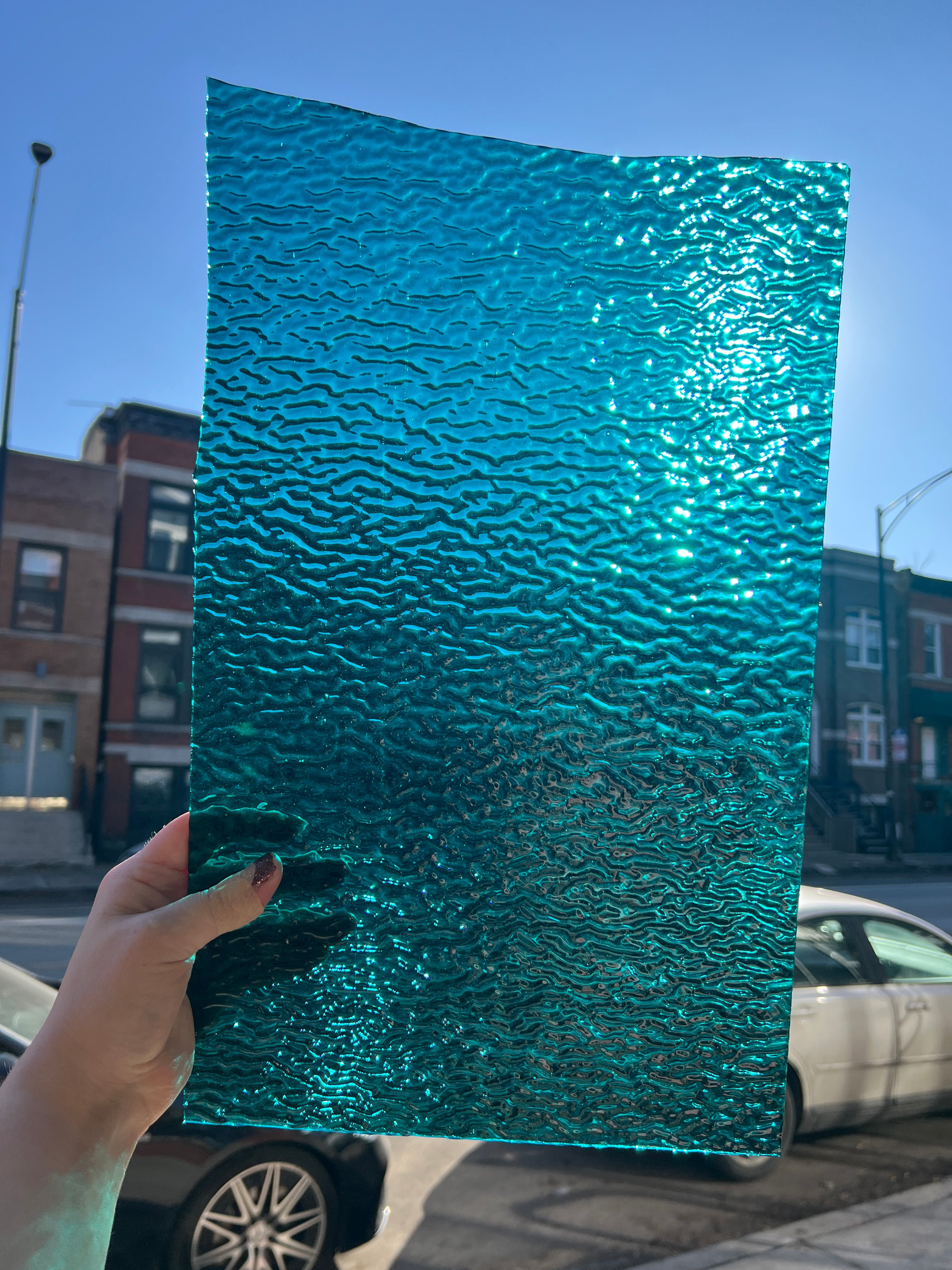 Person holding a textured teal stained glass piece outdoors with buildings and a car in the background