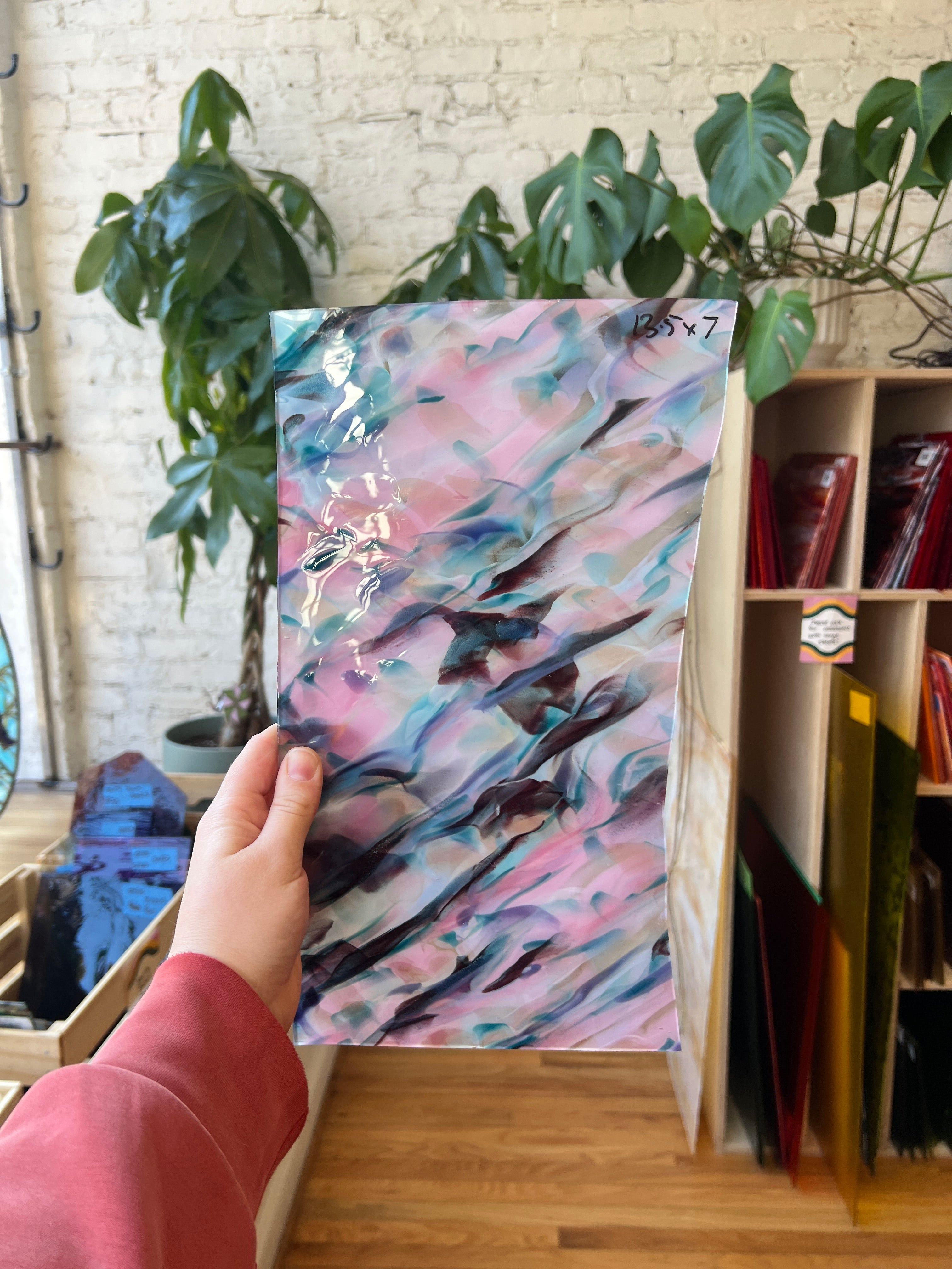 Hand holding a colorful abstract piece of stained glass in front of a plant and bookshelf.