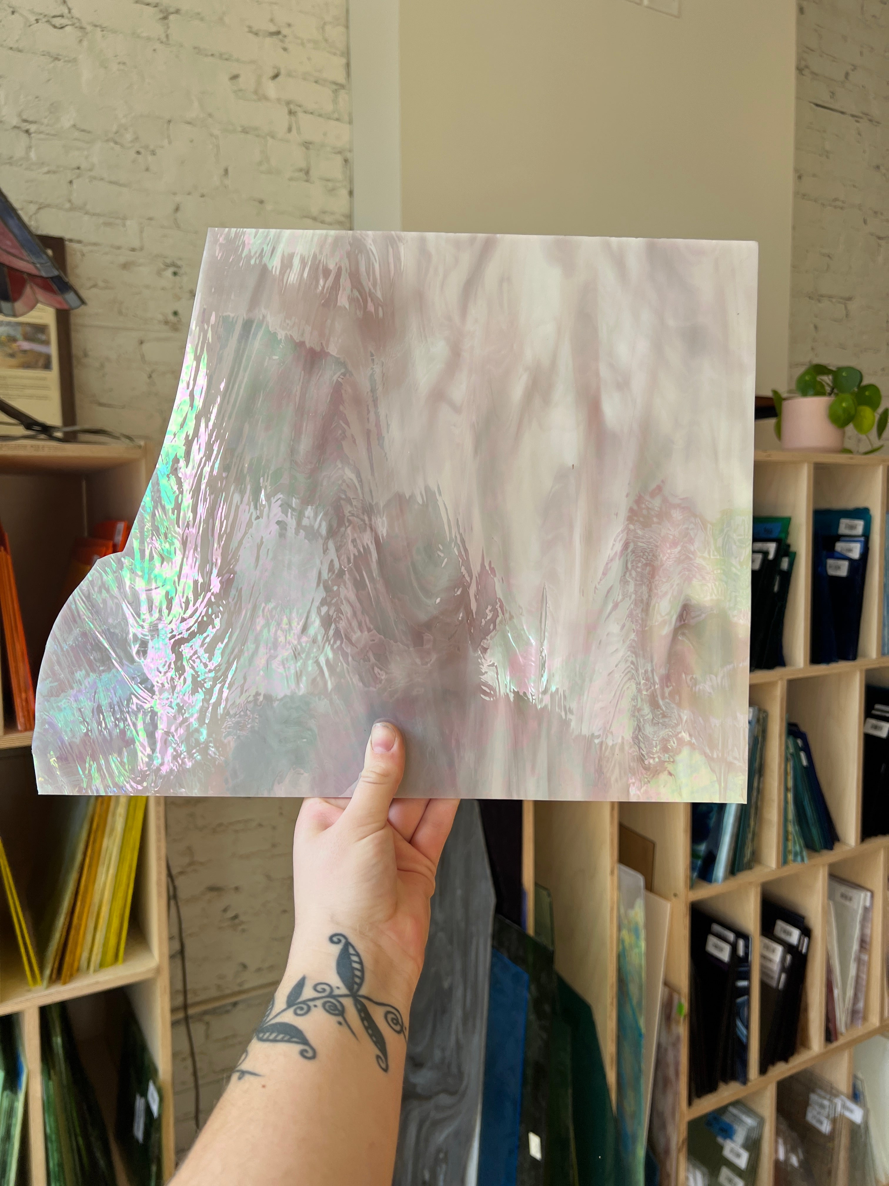 Hand holding a sheet of iridescent glass in an indoor setting with shelves in the background.