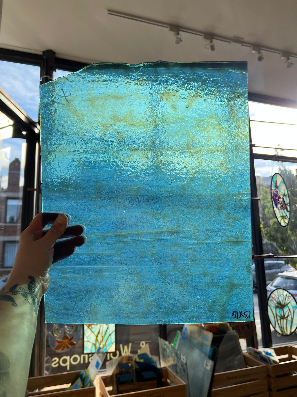 A hand holds up a large, textured square glass panel with a mottled blue and turquoise pattern, set against a storefront window background.