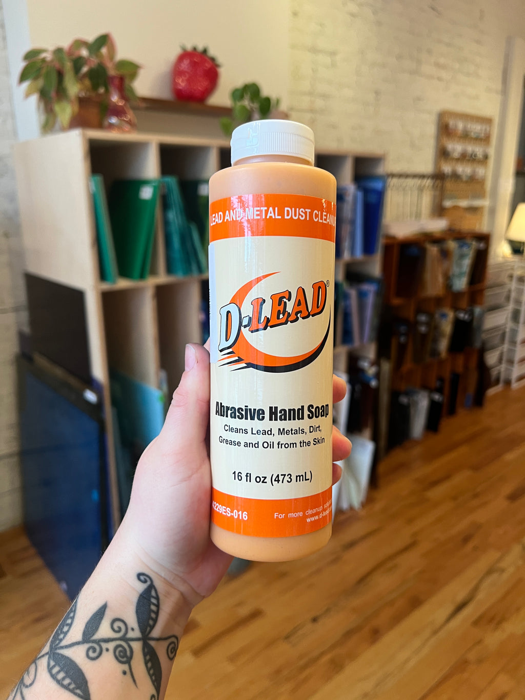 Hand holding a bottle of D Lead abrasive hand soap with a blurred indoor background.