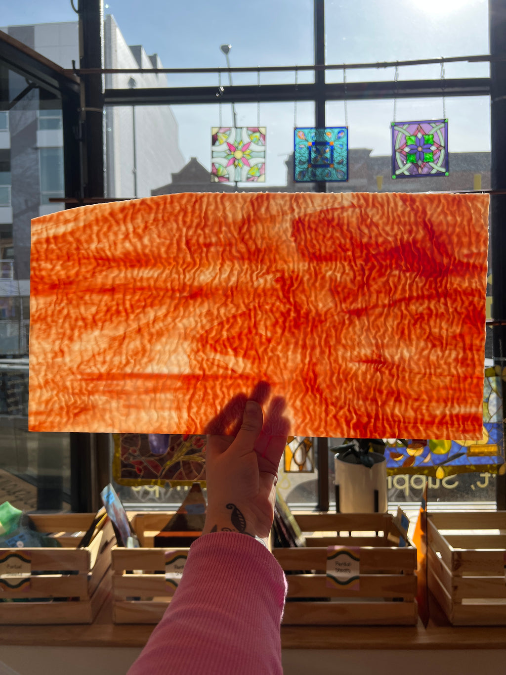 Person holding an orange textured piece of stained glass in front of a window with colorful artworks.