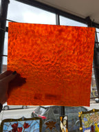 Hand holding an orange textured square of glass in front of a stained glass window
