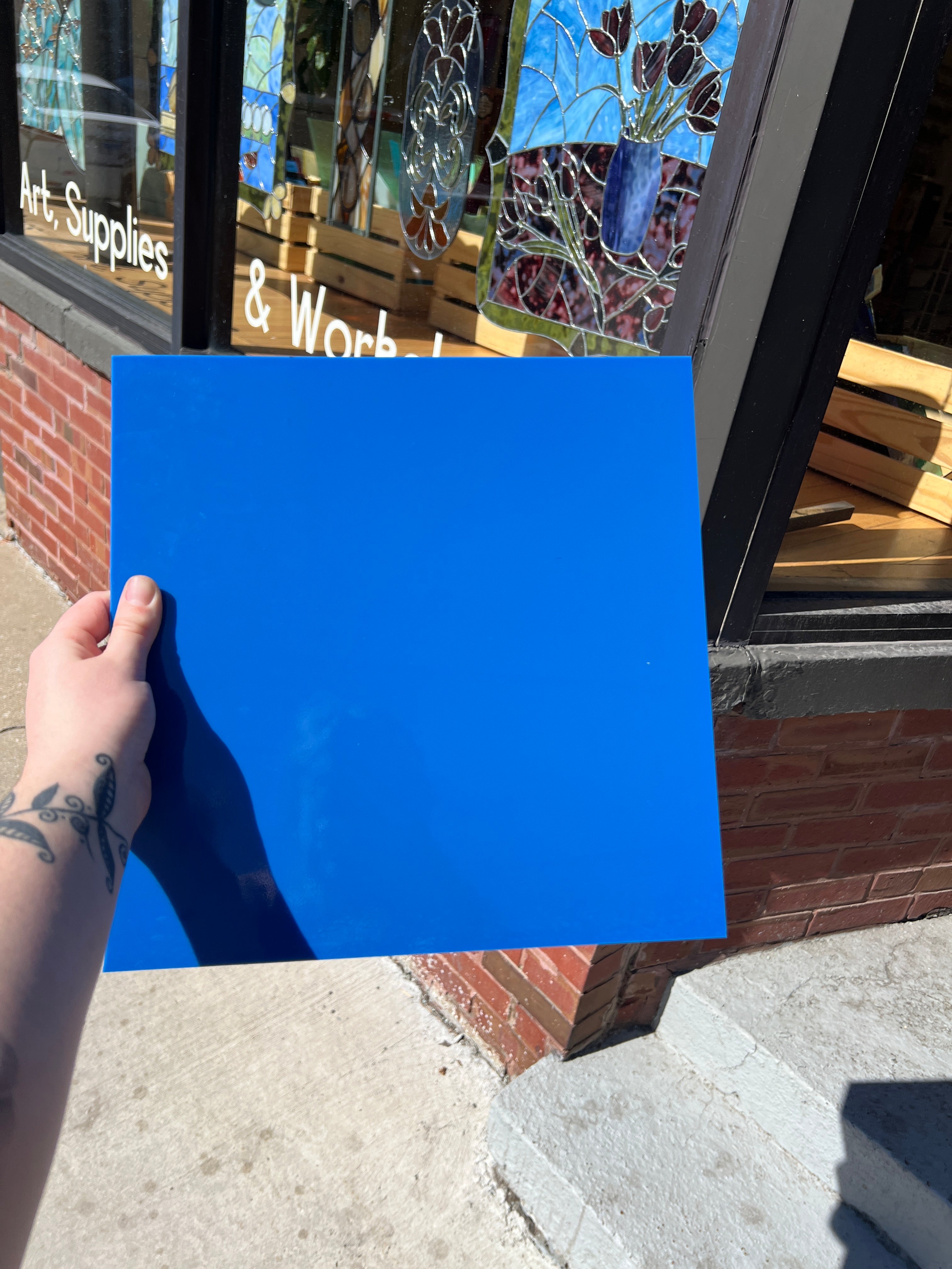 Hand holding a blue square of glass in front of a store window with stained glass displays.
