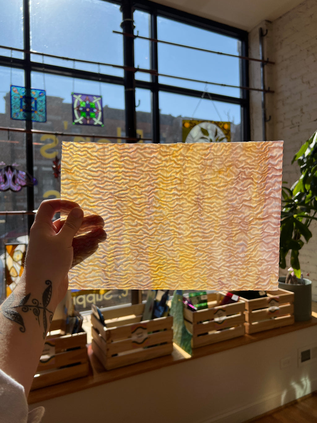 Person holding a yellow and tan textured piece of stained glass in front of a blurred sunny window. 