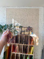 Hand holding a lightly textured peachy brown piece of glass in front of a colorful shelf.