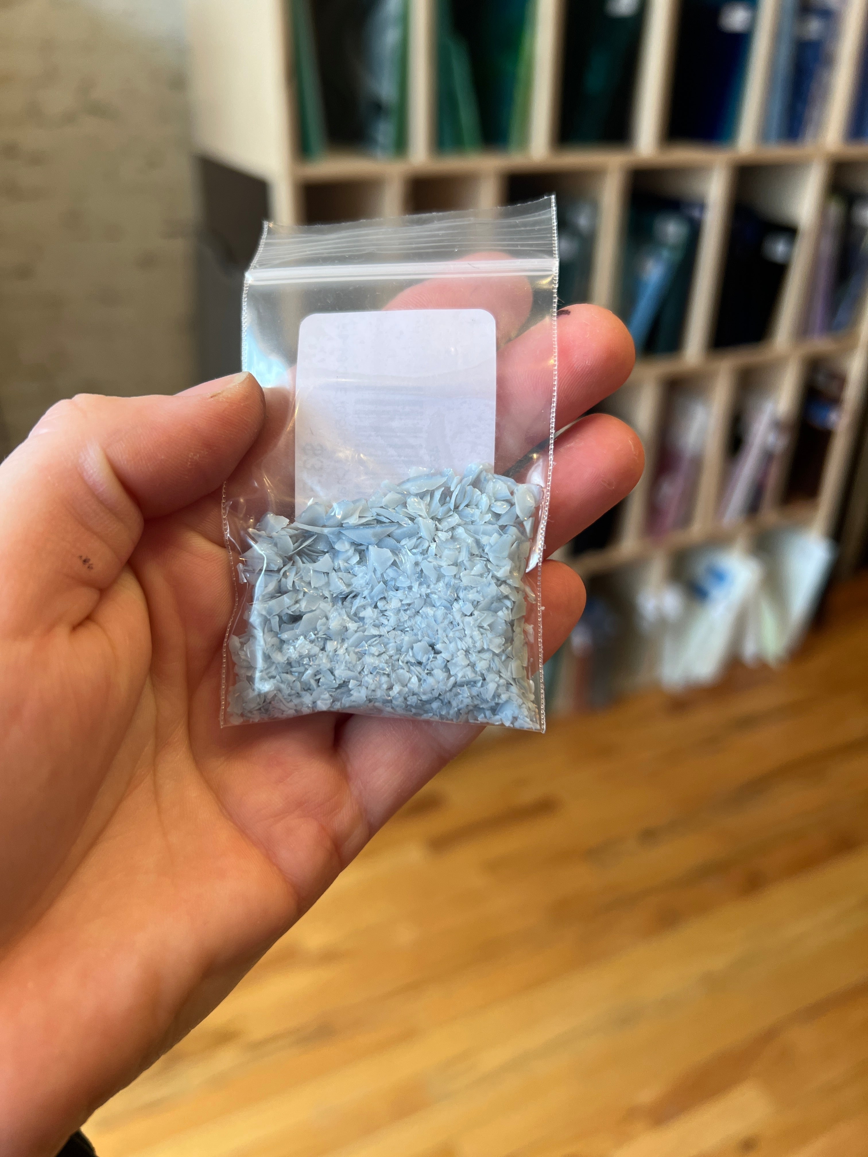 Hand holding a small clear plastic bag with blue gray frit crystals against a blurred background
