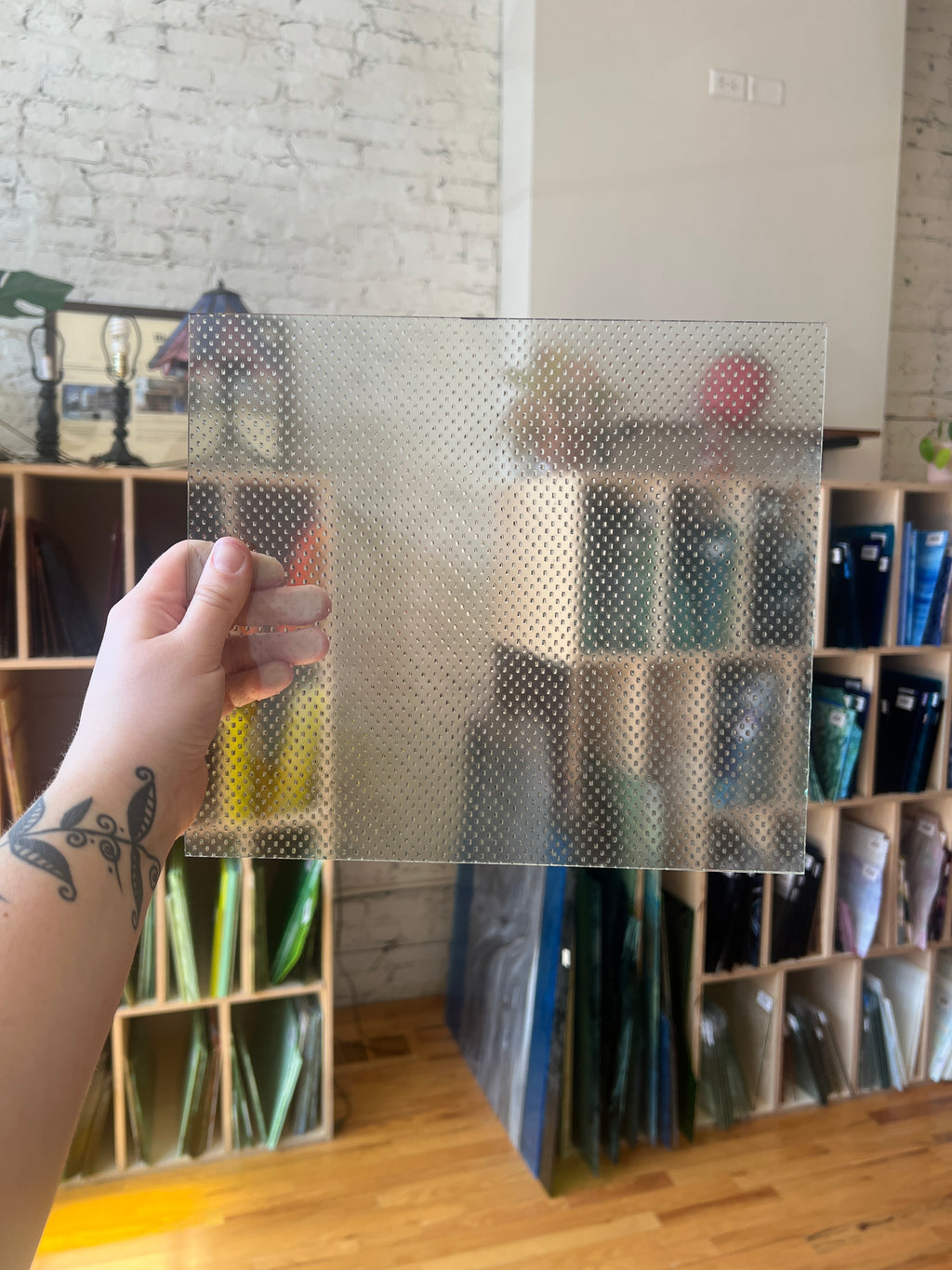 Hand holding a transparent textured sheet of glass against a background of shelves with various items.