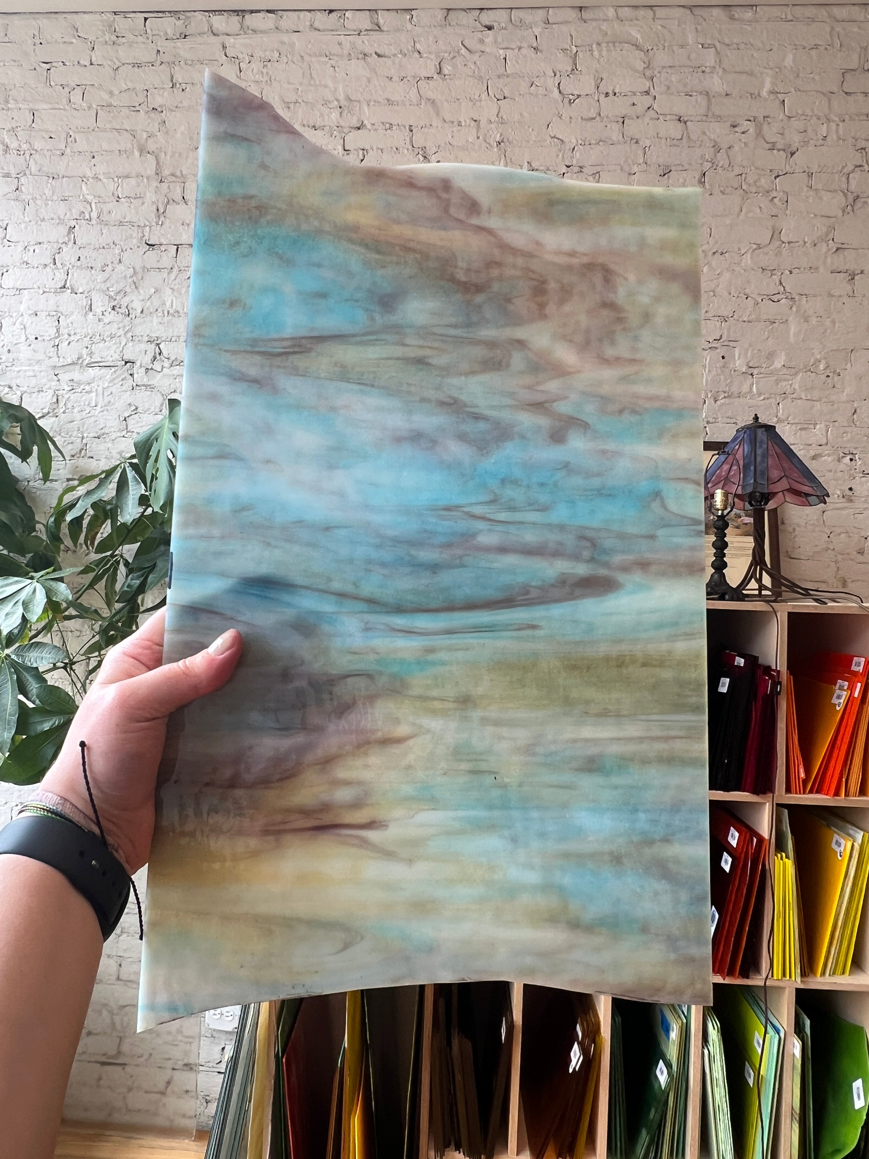 Hand holding a piece of stained glass with a watercolor-like pattern against a textured wall.