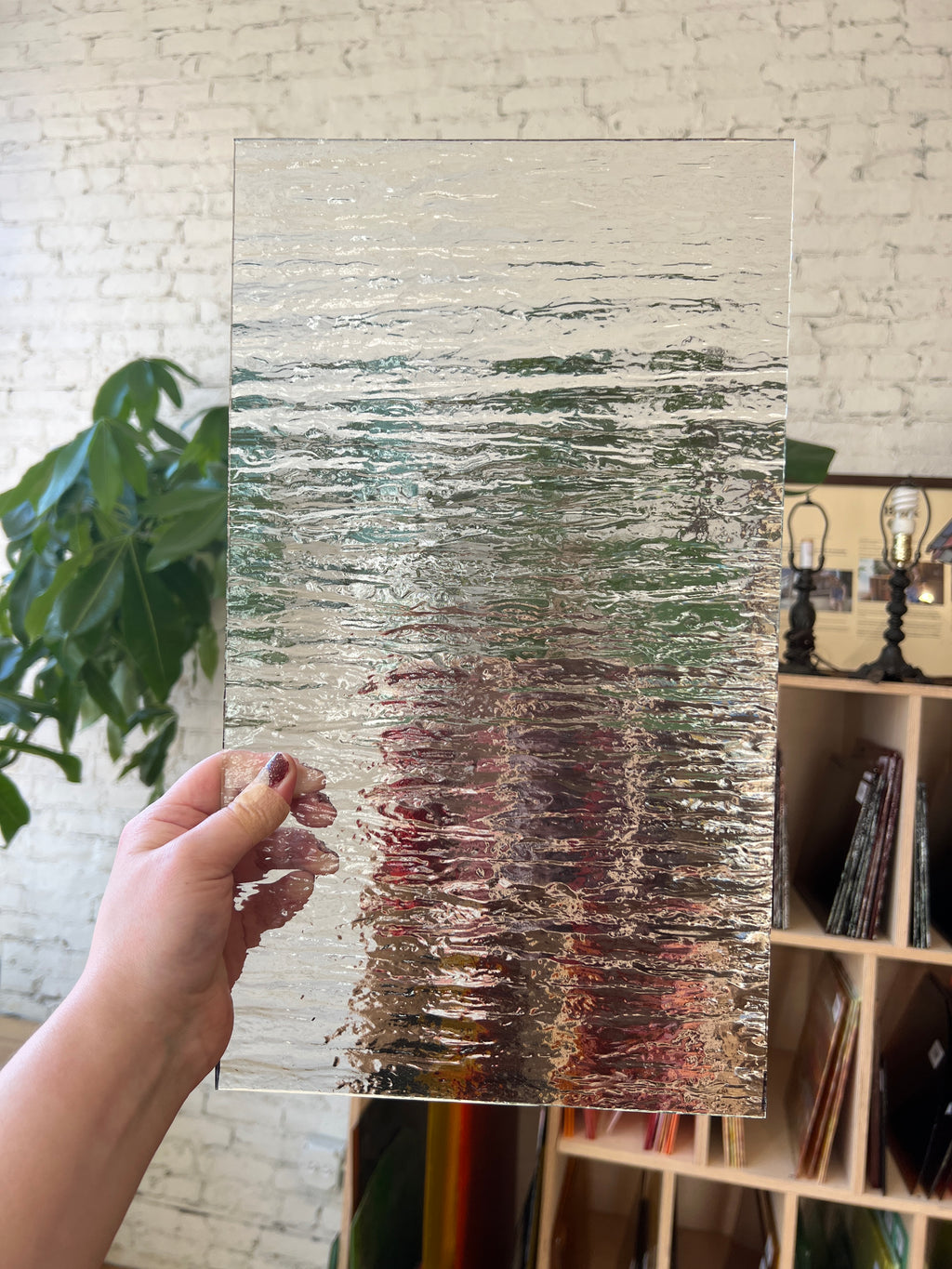Person holding a piece of clear textured glass in front of a blurred background of a shelf and brick wall. 