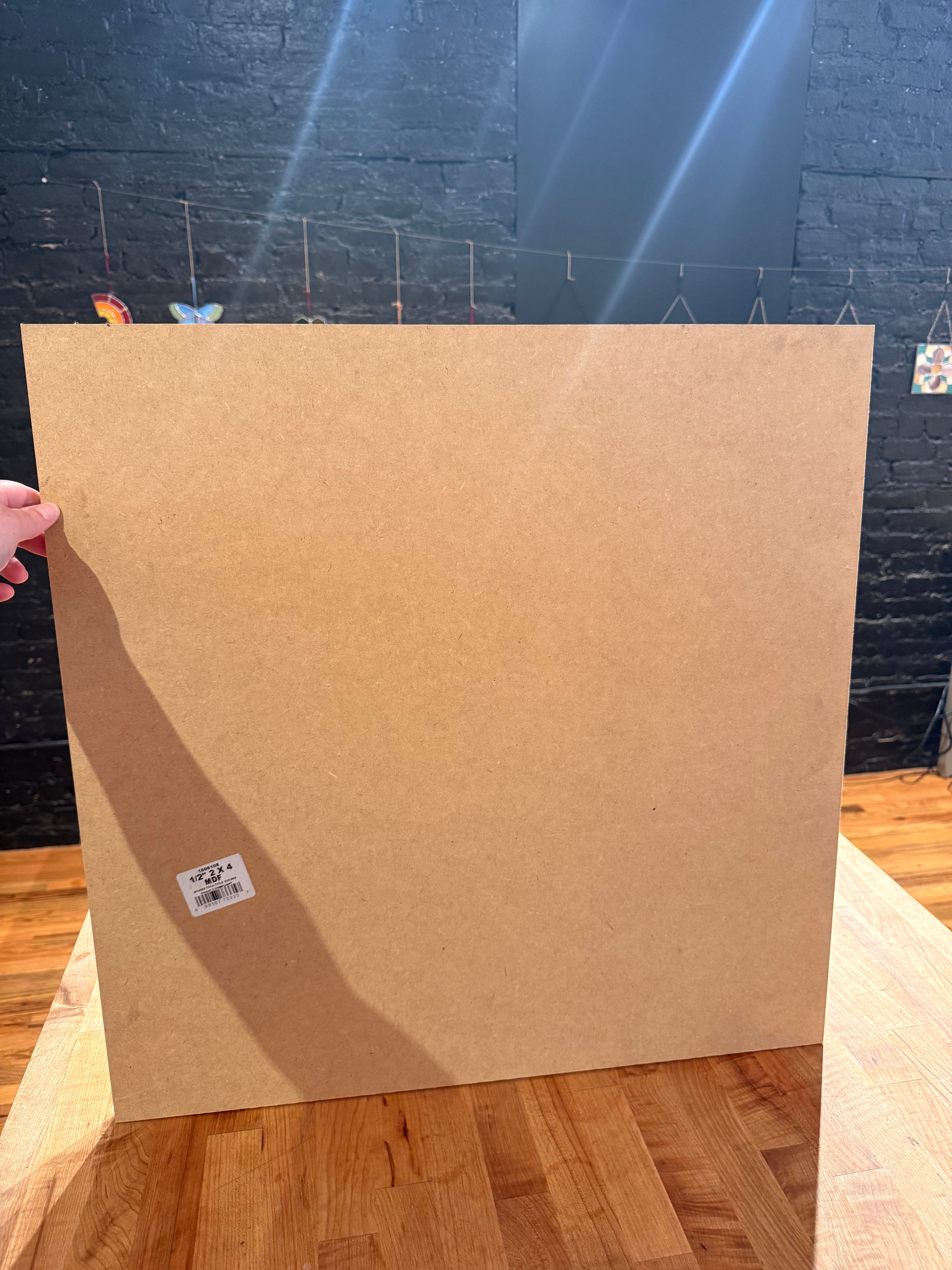 A large square piece of hemasote board held upright by a hand on a wooden table, with a dark brick wall in the background.