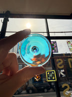 A hand holds a round, clear glass piece with a vibrant blue and iridescent rainbow reflection in front of a window with stained glass panels.