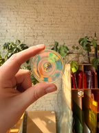 A hand holds a circular clear rainbow glass disc in front of a sunlit brick wall and indoor plants.
