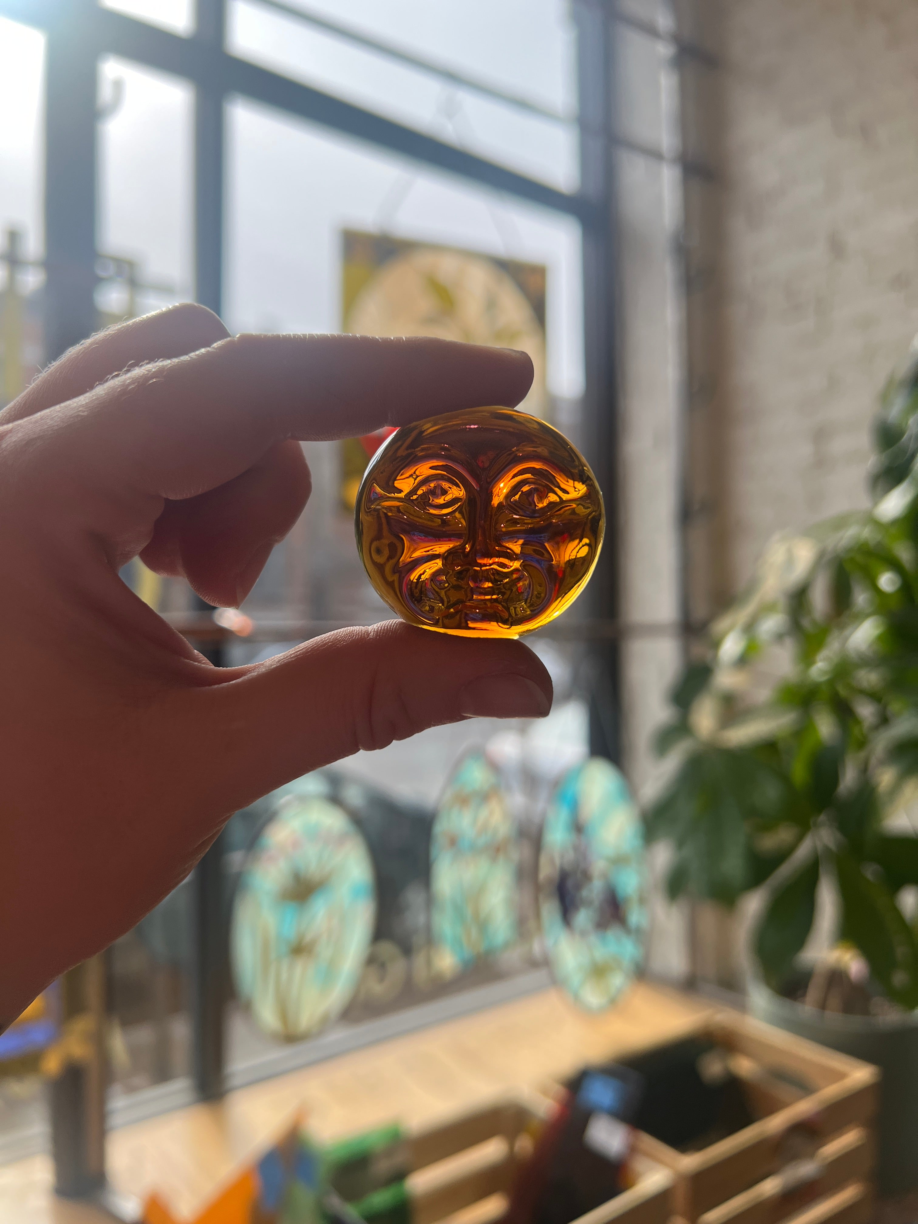 Hand holding a round, amber-colored glass jewel with intricate moon face designs against a blurred indoor background