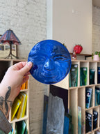 Hand holding a blue glass object with a textured surface, set against a background of art supplies.