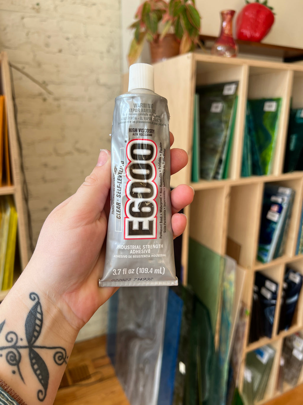 Hand holding a tube of E6000 in front of blurred shelves