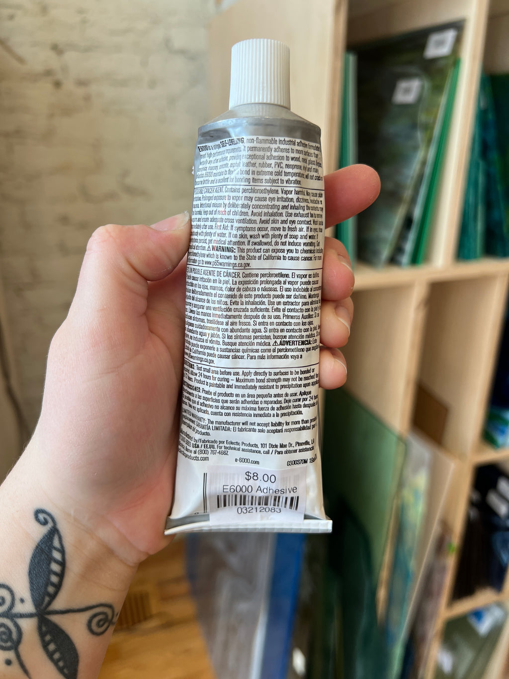 Hand holding up the back of a tube of E6000 in front of blurred shelves.