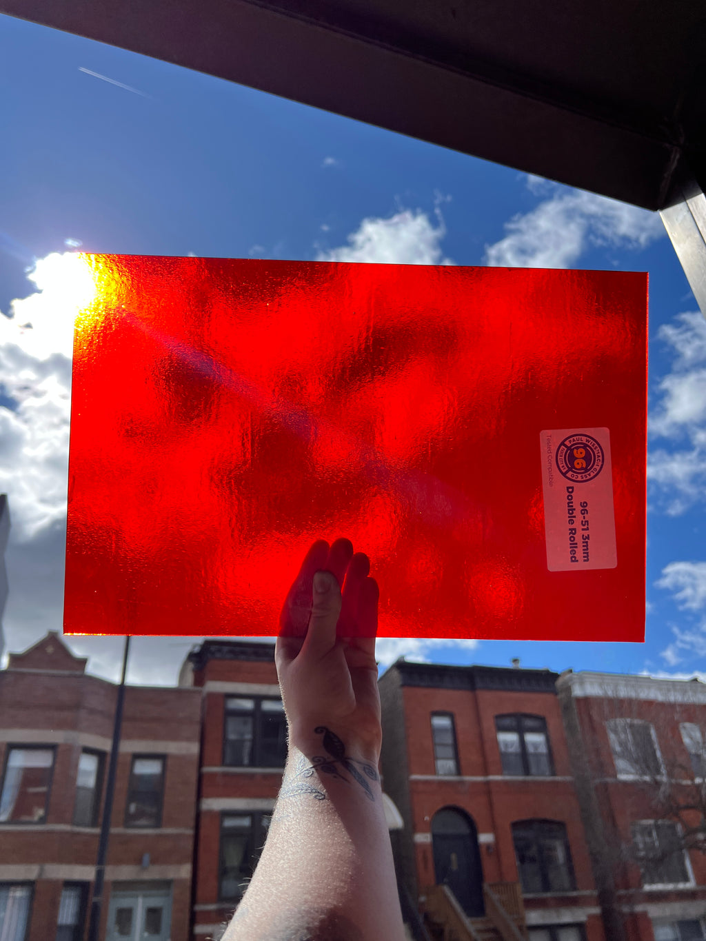 Red sheet of stained glass held up against a blue sky with buildings in the background