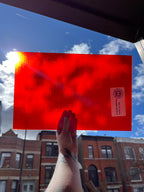 Red sheet of stained glass held up against a blue sky with buildings in the background
