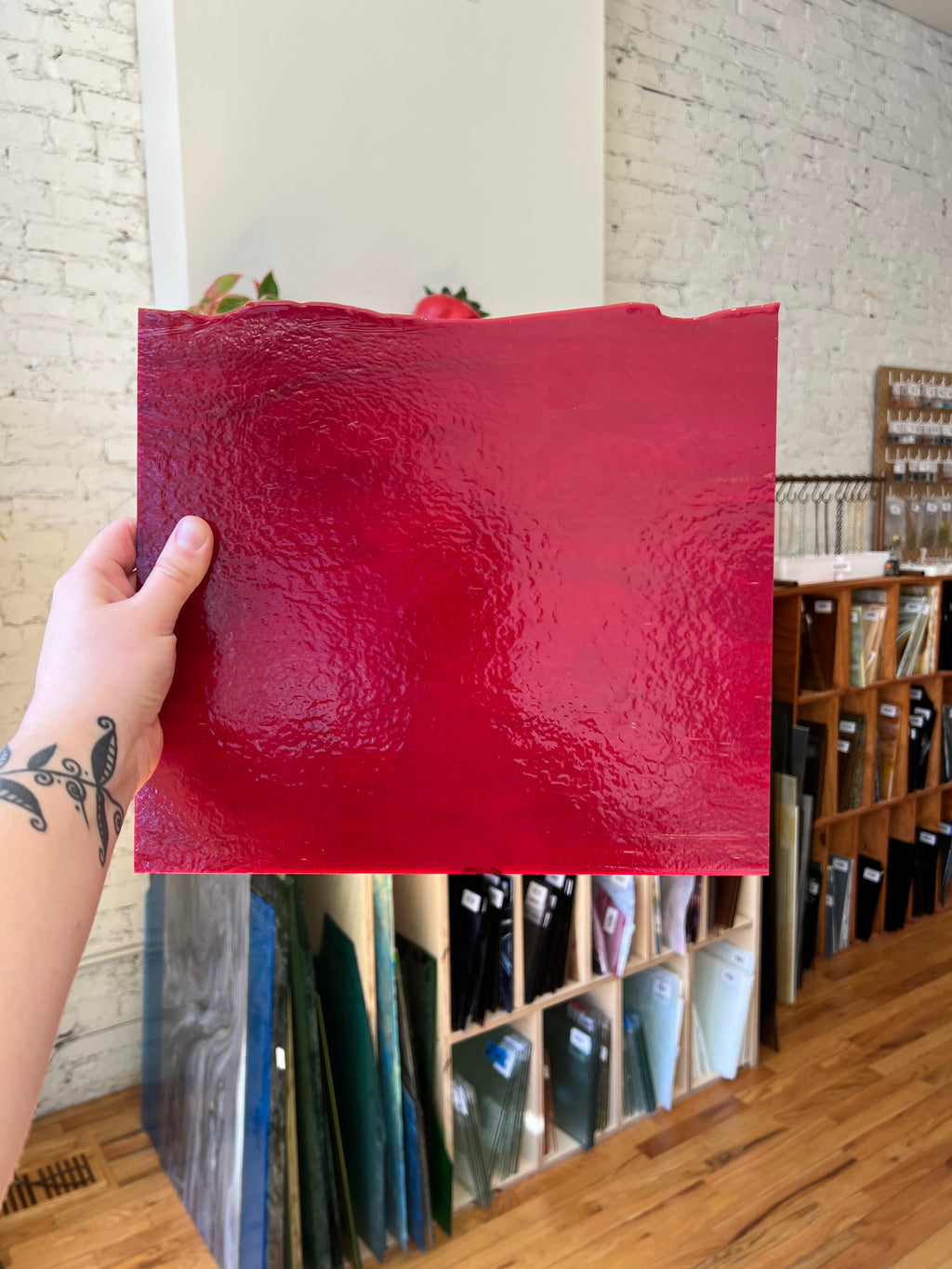 Red textured piece of glass held by a hand with tattoos, with a shelf in the background