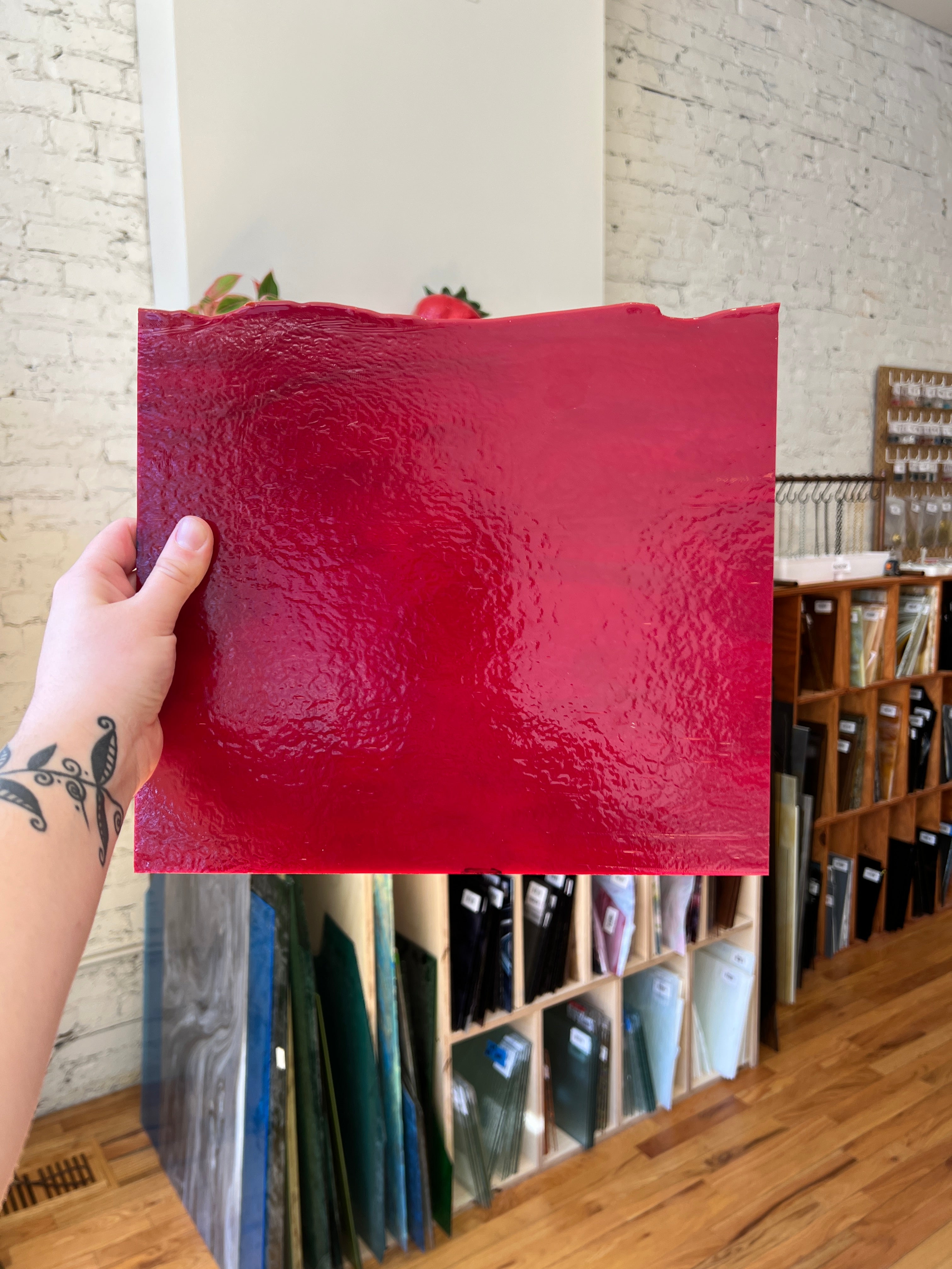 Red textured piece of glass held by a hand with tattoos, with a shelf in the background