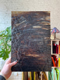 Brown sheet of stained glass held by a hand with a blurred background of colorful items