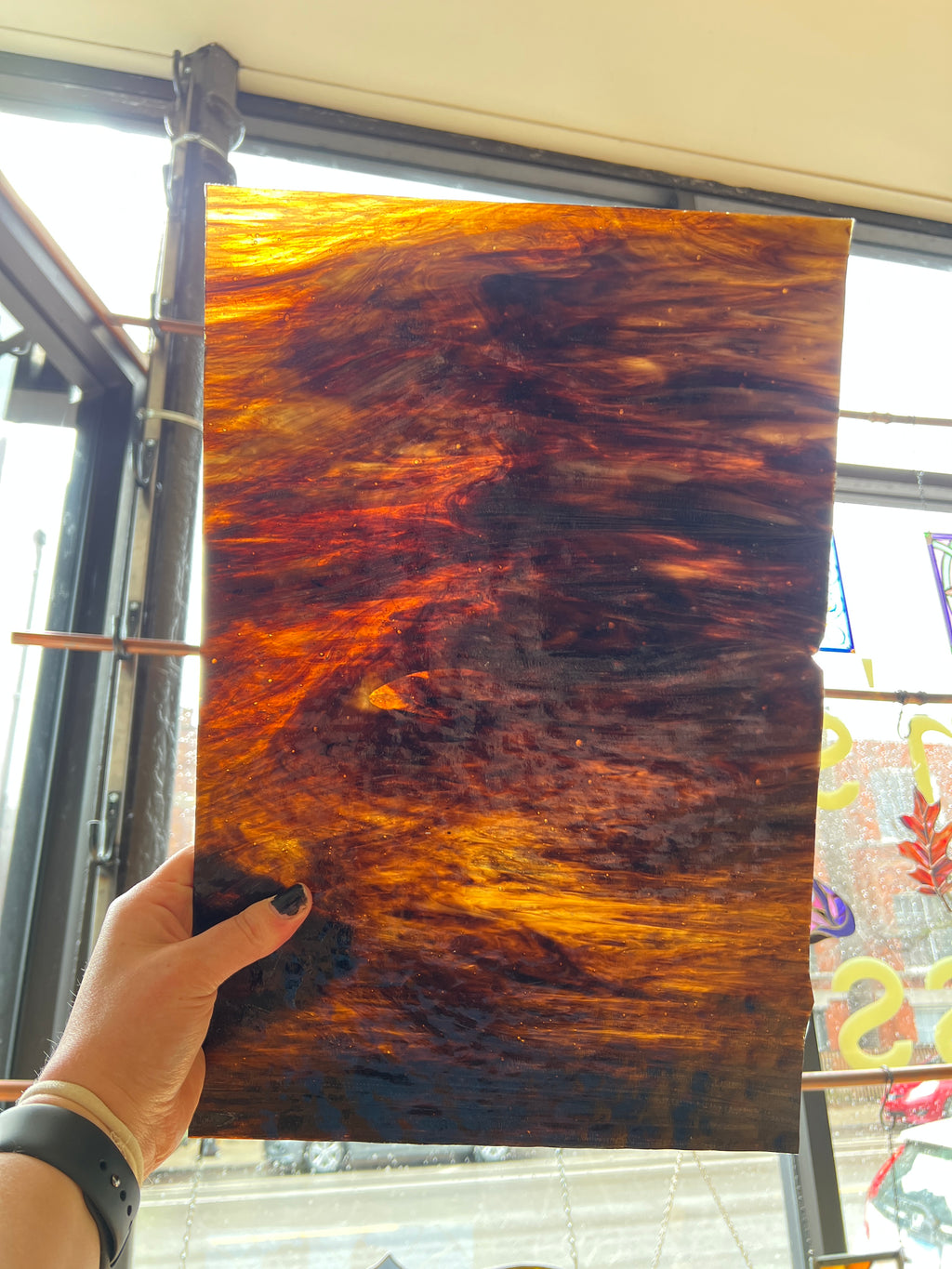 Hand holding a large piece of brown stained glass with a distinctive grain pattern in front of a window.