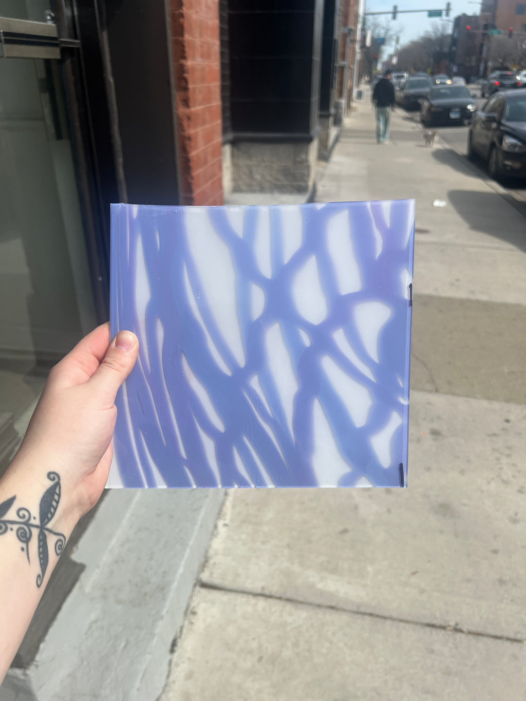Hand holding a blue and white patterned piece of stained glass on a city street.