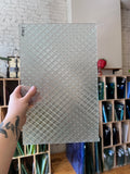 Hand holding a textured glass sheet with a pattern, with a blurred background of shelves.