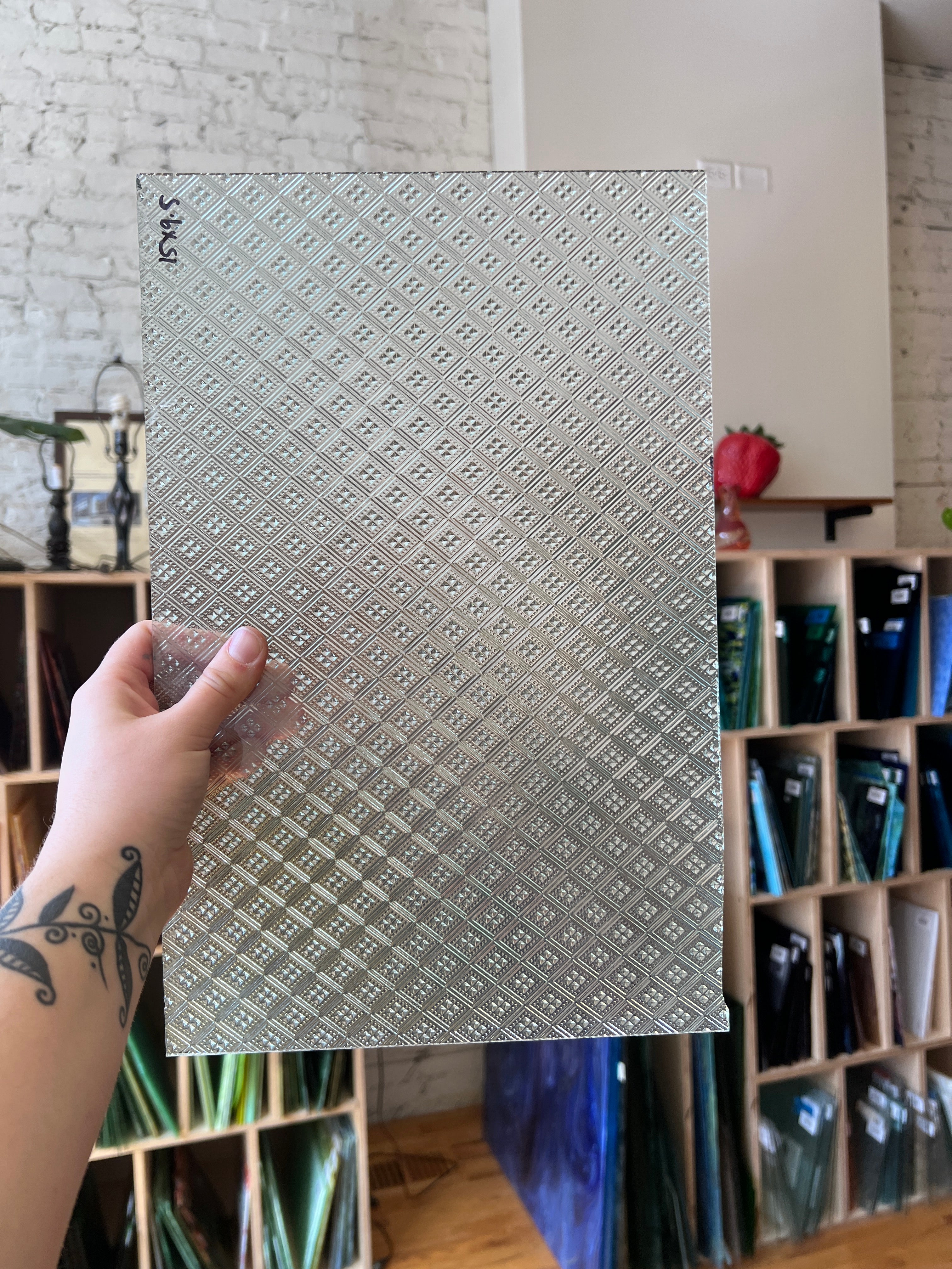 Hand holding a textured glass sheet with a pattern, with a blurred background of shelves.