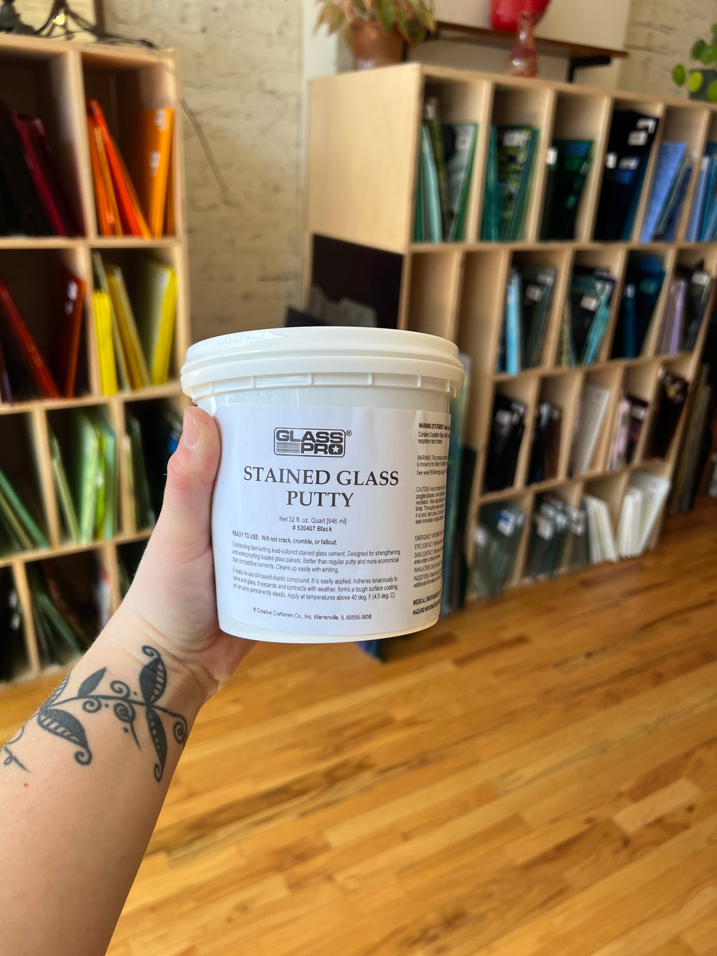 Hand holding a container of stained glass putty in front of a display case with various items.