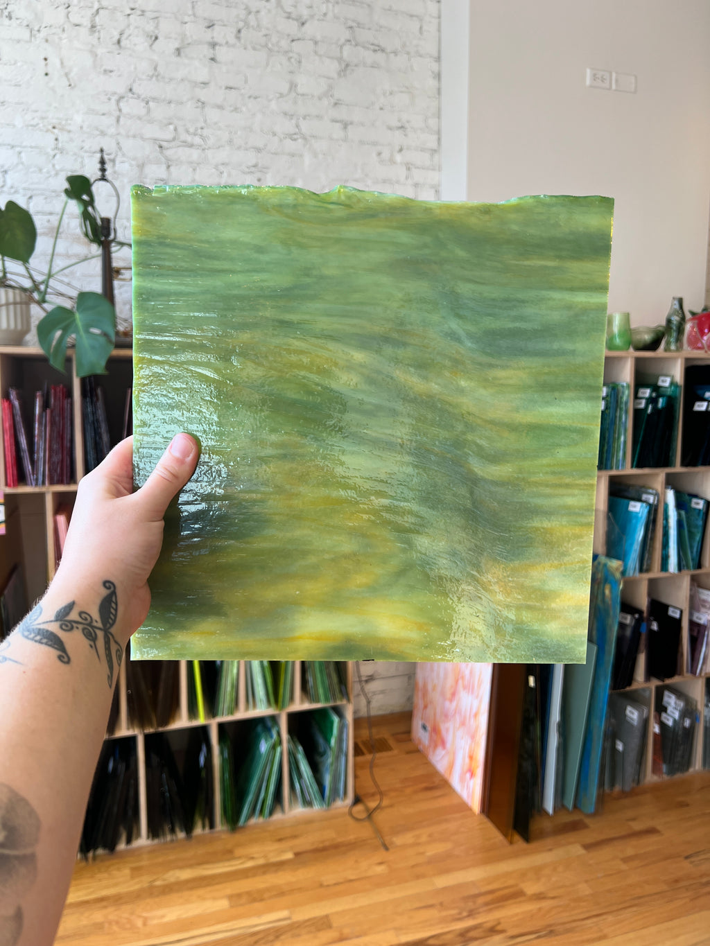 A hand holding a green and yellow streaky piece of stained glass in front of colorful shelves