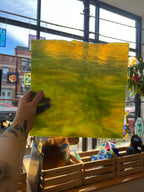 A hand holds a square glass art piece with a textured, rippled surface showing a gradient of yellow and green, set against a window view of a storefront.