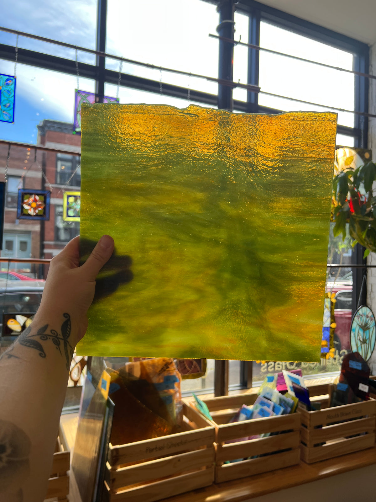 A hand holds a square glass art piece with a textured, rippled surface showing a gradient of yellow and green, set against a window view of a storefront.