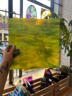 A hand holding a green and yellow streaky piece of stained glass in front of a city window