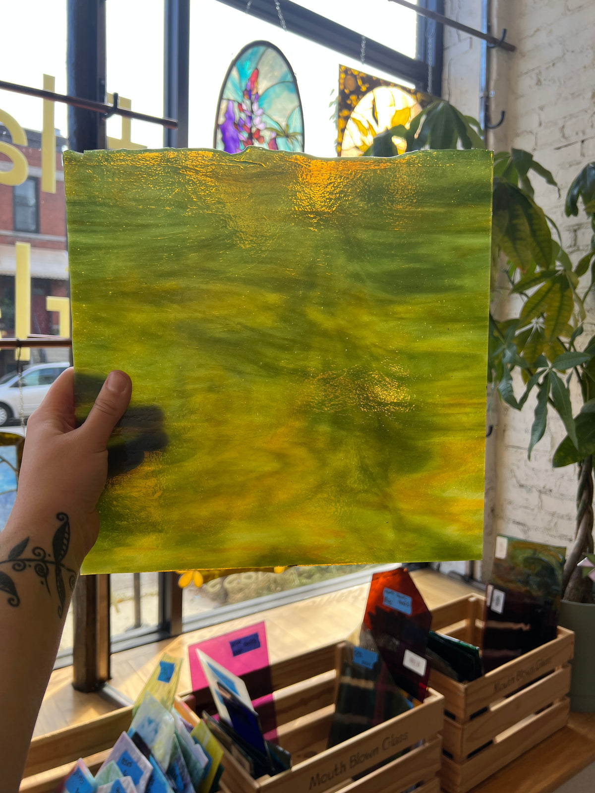 A hand holding a green and yellow streaky piece of stained glass in front of a city window