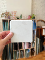A hand holding a blank, square, off-white stone tile or coaster in front of a blurred background of storage boxes and glassware.