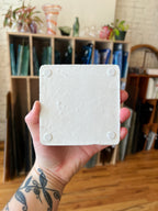 A hand with a black tattoo holds a square white textured tile or coaster in front of a blurred indoor background with shelves and wood flooring.