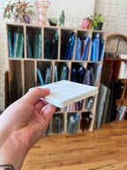 A hand holding a white rectangular coaster in front of a wooden display organizer filled with colorful glass panes.