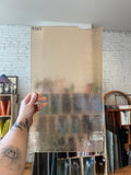 A hand holds a large rectangular glass panel with a textured, frosted appearance in an indoor setting with shelves and a white brick wall in the background.