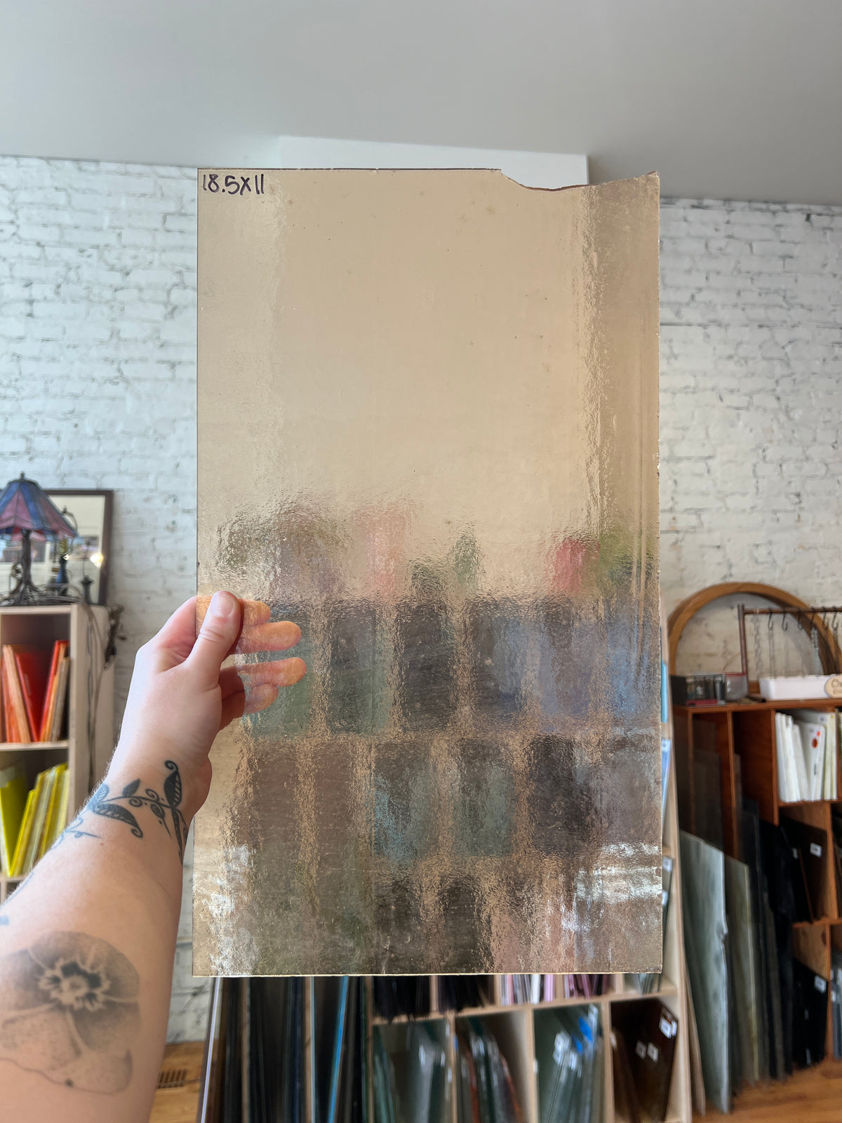 A hand holds a large rectangular glass panel with a textured, frosted appearance in an indoor setting with shelves and a white brick wall in the background.