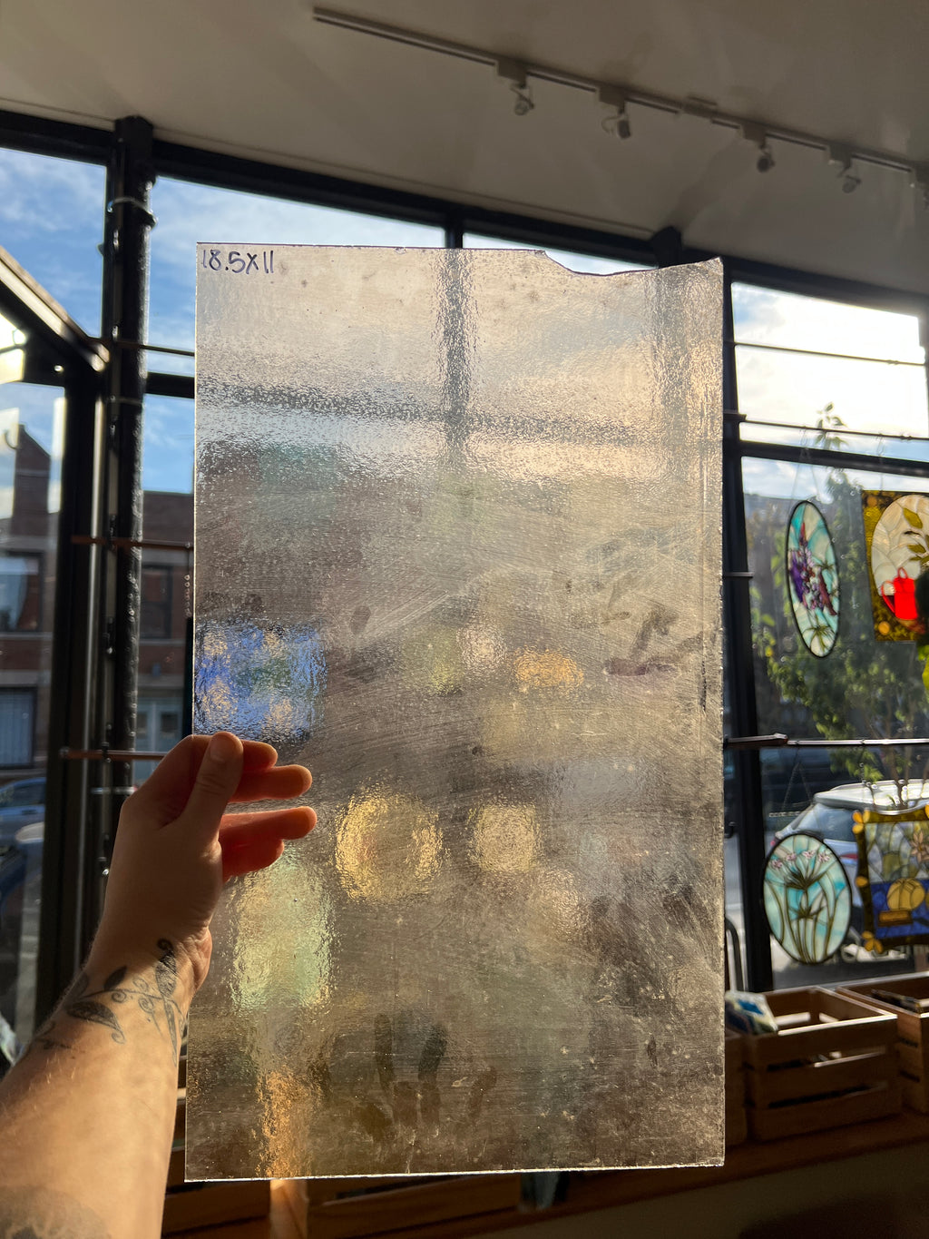 A hand holds a large rectangular sheet of textured, frosted translucent glass in front of a window with stained glass panels in the background.